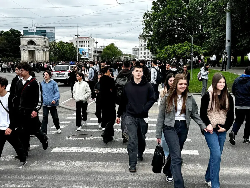 Crowds of people cross a city street in Chisinau, Moldova, with historic buildings and trees in the background.