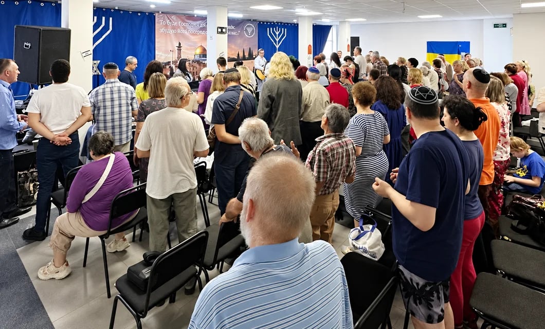 A large congregation gathered for worship in a hall decorated with menorah symbols and a Ukrainian flag.